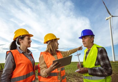 Üç mühendis rüzgar türbini çiftliğinde raporu tutuyor ve planı tartışıyorlar. Korunmak için floresan yelek ve kask giyiyorlar..