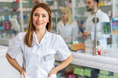 Smiling portrait of young, beautiful female pharmacist that is standing at the cash desk. Other pharmacists working in the background.
