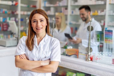 Smiling portrait of young, beautiful female pharmacist that is standing at the cash desk. Other pharmacists working in the background.