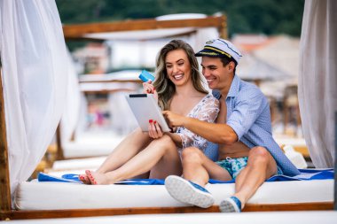 Genç ve güzel bir çift sahilde kartla internetten sipariş veriyor. Denizci şapkası takan erkek arkadaş..