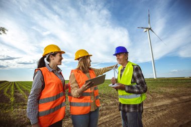 Üç mühendis rüzgar türbini çiftliğinde raporu tutuyor ve planı tartışıyorlar. Korunmak için floresan yelek ve kask giyiyorlar..