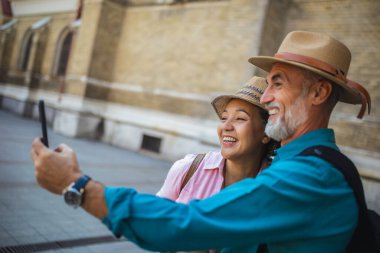 Yeni şehri keşfeden yaşlı evli bir çift. Merkezde selfie çekiyoruz. Şapka ve sırt çantası takıyorlar..