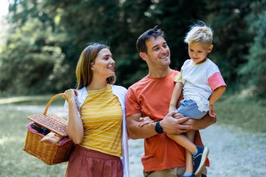 Üç kişilik aile parkta pikniğe gidiyor. Sepet, battaniye ve yiyecek tutuyorlar..