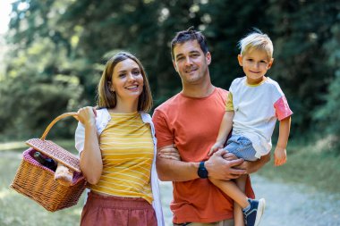 Üç kişilik aile parkta pikniğe gidiyor. Sepet, battaniye ve yiyecek tutuyorlar..