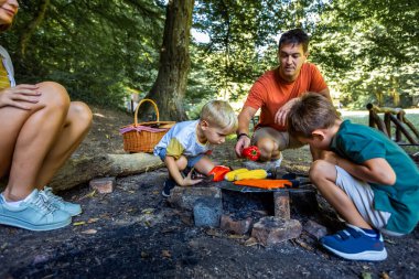Dört kişilik bir aile ormanda öğle yemeği molası veriyor. Oğullar babalarına et ve sebze ızgarasında yardım ediyorlar..