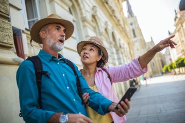Yaşlı evli çift yeni şehri keşfediyor. Navigasyon ve harita için telefonlarını kullanıyorlar. Şapka ve sırt çantası takıyorlar..