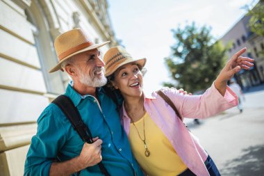 Şehri keşfeden olgun bir çift. Birbirlerine sarılıyorlar. Şapka ve sırt çantası takıyorlar..