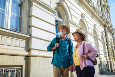 Yaşlı evli çift el ele tutuşup yeni şehirde yürüyor. Sırt çantaları ve şapkaları var. Keşif ve seyahat konsepti.