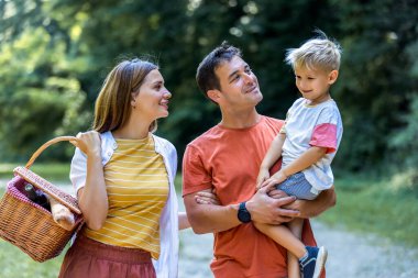 Üç kişilik aile parkta pikniğe gidiyor. Sepet, battaniye ve yiyecek tutuyorlar..