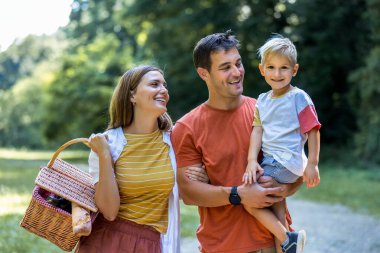 Üç kişilik aile parkta pikniğe gidiyor. Sepet, battaniye ve yiyecek tutuyorlar..