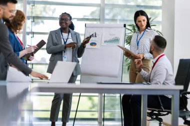 İki iş kadını bir ofiste diğer iş arkadaşlarıyla konuşuyor. Grafikler, diyagramlar ve istatistikler gösteriliyor.