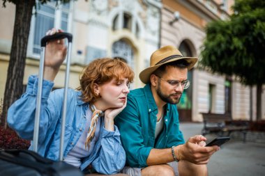 Yorgun çift şehir merkezinde oturdu. Harita arıyorlar çünkü keşfettikleri şehirde kayboldular. Bavulları ve sırt çantaları var..
