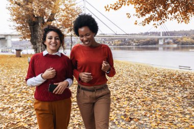 İki gülümseyen genç bayan arkadaş ormanda gölün yanında kırmızı kazaklarla koşuyorlar.
