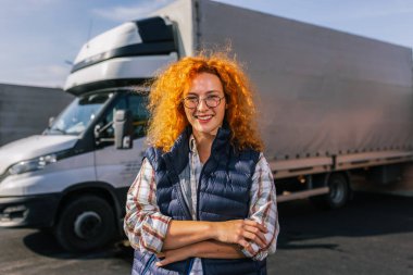 Kızıl saçlı kadın kamyon şoförünün portresi. Uzun aracının önünde duruyor..
