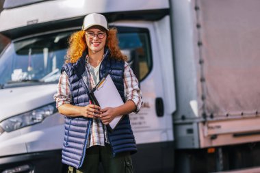 Kızıl saçlı kadın kamyon şoförü elinde bir kağıtla kamyonun önünde duruyor ve bir şeyler yazıyor. Ulaştırma sektörü teması.