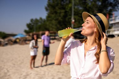 Güzel genç bir kadın sahilde kokteyl içerken arkadaşları onun arkasında oynaşıyor..