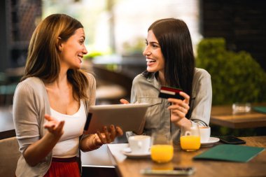 İnternetten alışveriş yapmak için en iyi iki kız arkadaş kafede buluştu. Tablet ve kredi kartı kullanıyorlar. Arkadaşlar takılır, kahve ve portakal suyu içer..