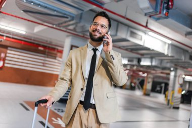 Modern iş adamı otoparkta bavulla yürüyor. İş gezisine gidiyorum. Telefonunu kullanarak..