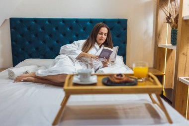 Otel odasında bornozlu, kitap okuyan güzel bir kız..