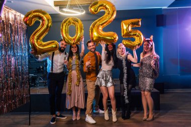 2025 yılbaşı arifesini kutlayan bir grup arkadaş. Altın balon numaralarını tutuyorlar..