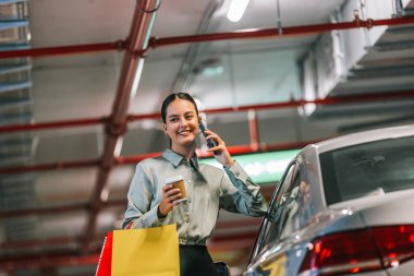 Young woman standing by her car with shopping bags using her phone and drinking coffee. clipart