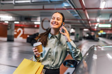 Arabasının yanında duran genç bir kadın alışveriş torbalarıyla telefonunu kullanıyor ve kahve içiyor..