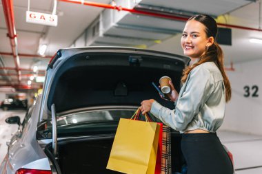 Young woman unlocking trunk of her car to put bags after shopping. clipart