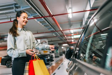 Young woman with shopping bags unlocking her car with keys in underground garage. clipart