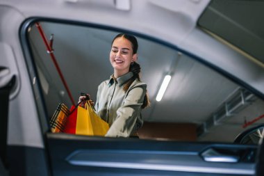 Young woman with shopping bags unlocking her car with keys in underground garage. clipart