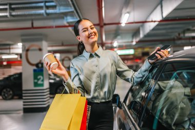 Young woman standing by her car with shopping bags using her phone and drinking coffee. clipart