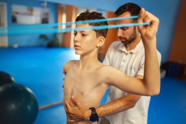 Küçük çocuğun terapi amaçlı kinezi terapisi. Egzersiz bantlarını çekerken omurgasını düzeltiyor. Bir kayropraktör ona yardım ediyor..