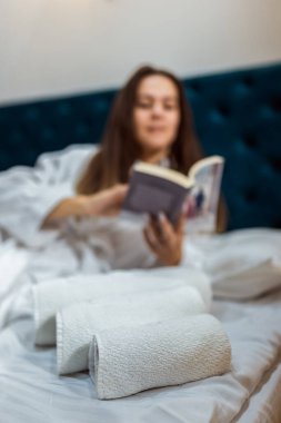 Yatakta havlulara yakın çekim. Arkalarında bornozlu kitap okuyan bir kız var..