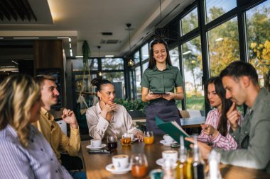 Garsondan yemek sipariş eden bir grup insan. Garson tablete emirler yazıyor. Garson onlara yardım ederken kararsız insanlar menüyü okuyor..