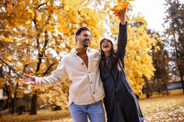 Genç mutlu çift parkta sarılıyor, yürüyor ve yaprak fırlatıyor. Güneşli sonbahar gününde Renkli park.