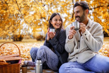 Genç mutlu çift renkli bir sonbahar parkında çimenlerde piknik yapıyor. Termostan kahve içiyorlar..