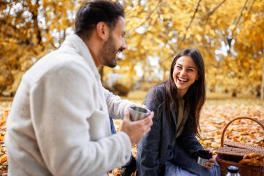 Genç mutlu çift renkli bir sonbahar parkında çimenlerde piknik yapıyor. Termostan kahve içiyorlar..