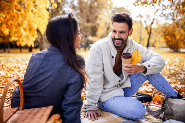 Genç mutlu çift renkli bir sonbahar parkında çimenlerde piknik yapıyor. Termostan kahve içiyorlar..