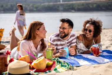 Üç arkadaş sahildeki piknikte renkli sağlıklı meyve suyu içerler..