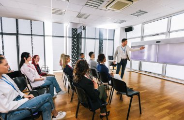 Erkek CEO, çok ırklı bir grup meslektaşın önünde duruyor. Üzerinde akıllı tahta ve grafikler olan çokluortam sınıfı.