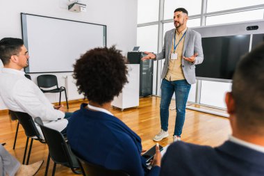 Erkek CEO, çok ırklı bir grup meslektaşın önünde duruyor. Üzerinde akıllı tahta ve grafikler olan çokluortam sınıfı.