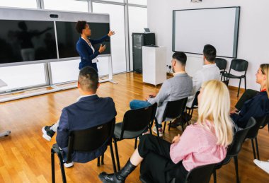 Afrikalı Amerikalı kadın CEO çok ırklı bir grup meslektaşın önünde duruyor. Üzerinde akıllı tahta ve grafikler olan çokluortam sınıfı.