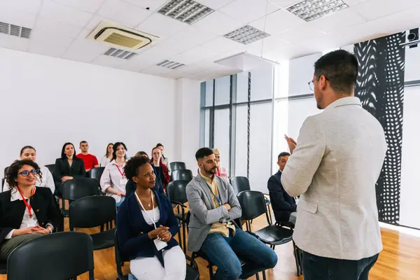 Erkek CEO, çok ırklı bir grup meslektaşın önünde duruyor. Üzerinde akıllı tahta ve grafikler olan çokluortam sınıfı.