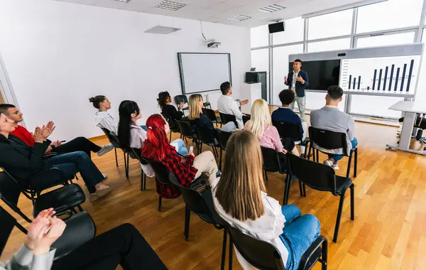 Asyalı erkek CEO, çok ırklı bir grup meslektaşın önünde duruyor. Üzerinde akıllı tahta ve grafikler olan çokluortam sınıfı.