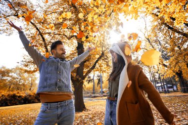 Genç mutlu çift parkta sarılıyor, yürüyor ve yaprak fırlatıyor. Güneşli sonbahar gününde Renkli park.