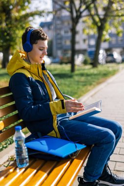 Parktaki ahşap bir bankta oturan, günlük kıyafetler giymiş, kulaklarında kulaklık olan bir çocuk. Elinde açık bir kitap tutuyor, okumaya dalmış.