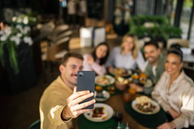 Bir grup arkadaş birlikte bir restoranda oturmuş, gülümsüyor ve akıllı telefonlarıyla selfie çekiyorlar..