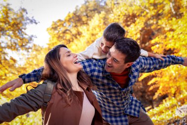 Gülümseyen baba, sonbahar parkında küçük oğlunu sırtında taşır. Annem gülümsüyor. Parktaki aile.