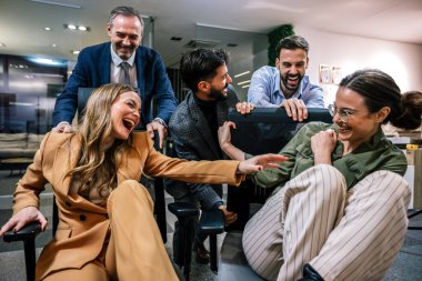 Modern bir ofiste, iş adamlarının dostça bir rekabete girdiği eğlenceli bir sahne. Resimde, meslektaşlar şakacı bir şekilde birbirlerini ofis sandalyelerinde itiyorlar.