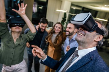 Modern bir ofiste VR kulaklık takarak ayakta durup eğlenen bir grup profesyonel.