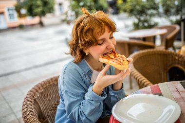 Genç kızıl kız bir dilim lezzetli pizzayı ısırıyor..